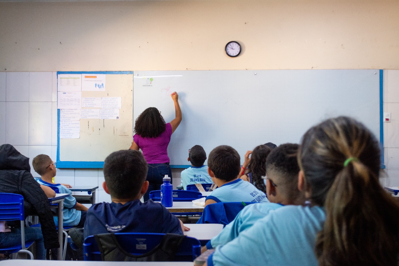 Imagem de uma sala de aula com uma professora escrevendo em um quadro branco